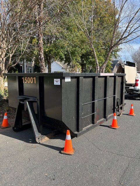 Large black dumpster with number 15001, surrounded by orange cones, placed on a suburban street.