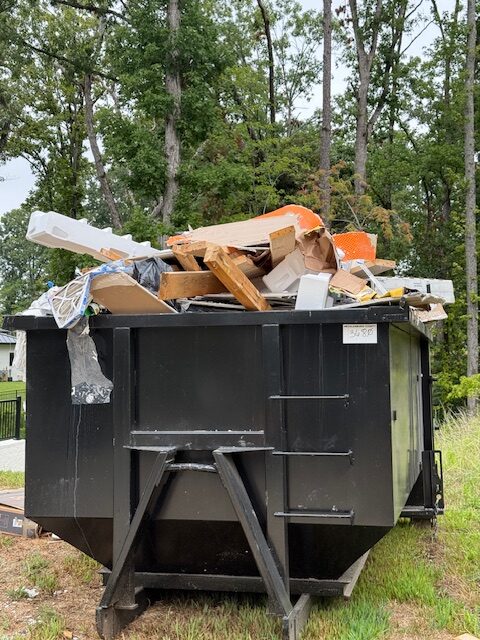 Dumpster filled with assorted debris including wood, cardboard, and plastic, set against a backdrop of tall, leafy trees and a cloudy sky.